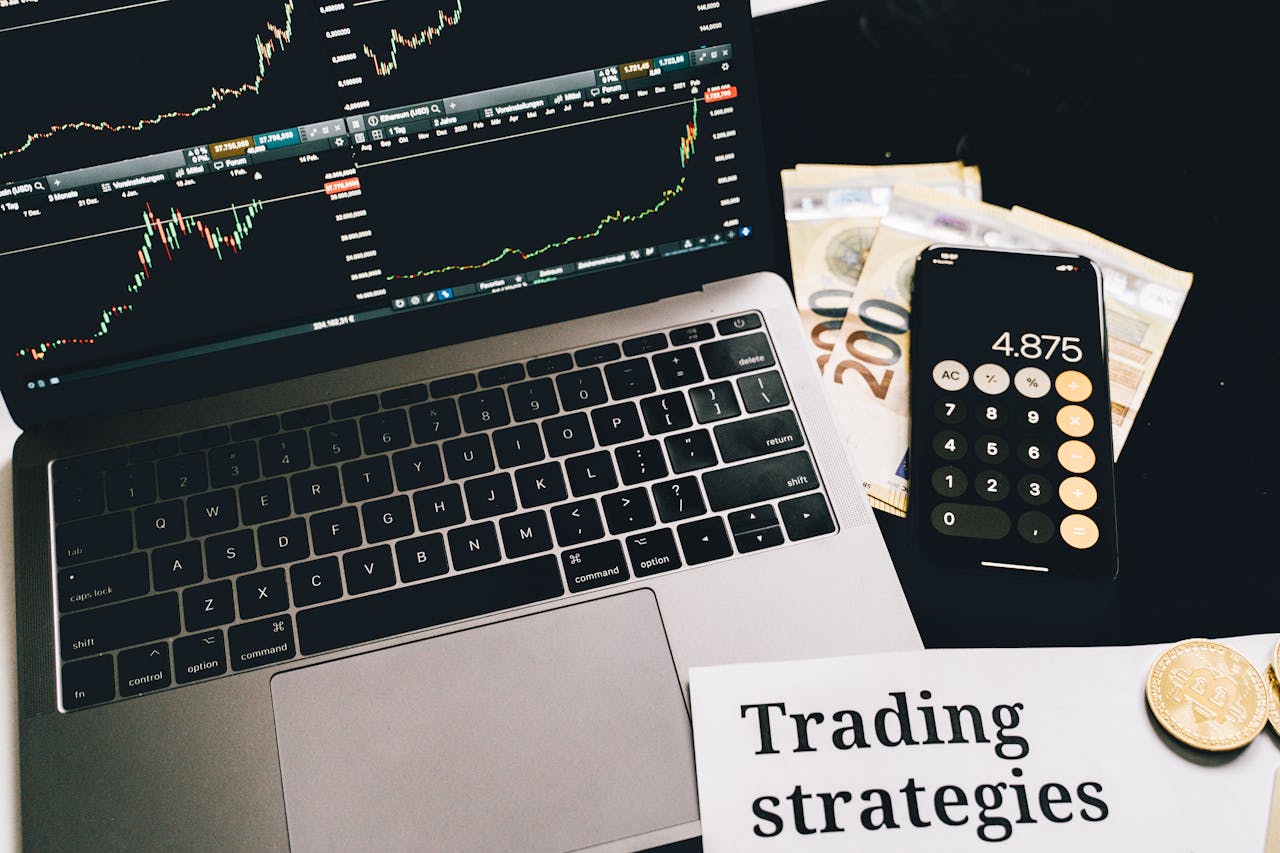 about-02 A laptop showing stock market graphs with a smartphone calculator, euro bills, coins, and trading strategies paper.