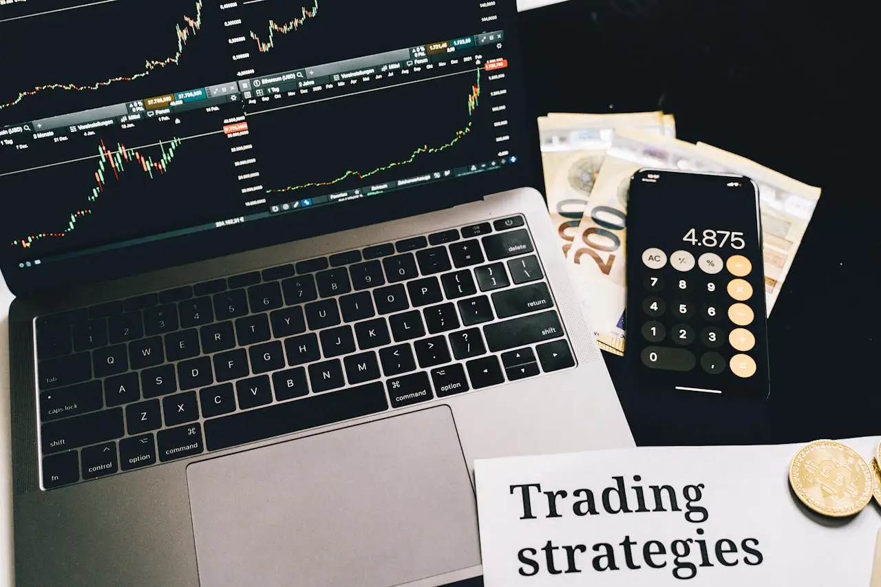 about-01 A laptop showing stock market graphs with a smartphone calculator, euro bills, coins, and trading strategies paper.